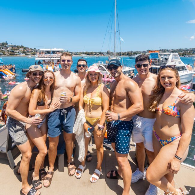 A group of eight young adults are posing together on a dock by the water, smiling and dressed in swimwear. Boats and water can be seen in the background under a clear, blue sky. The mood appears festive and summery, with laughter echoing across the lake.