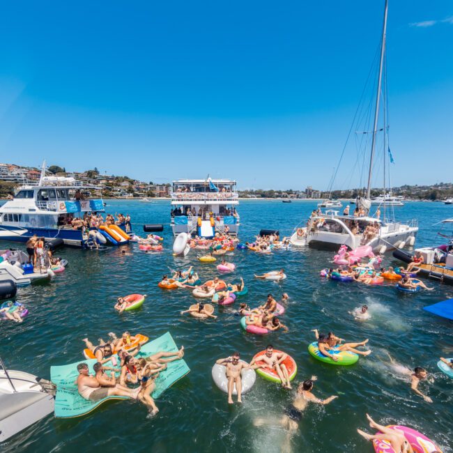 A large group of people enjoying a sunny day on the water, floating on inflatable tubes and lounge mats near anchored boats. The scene is festive with clear blue skies and bright sunlight, showcasing a vibrant gathering at sea. A yacht party adds to the lively atmosphere in the background.