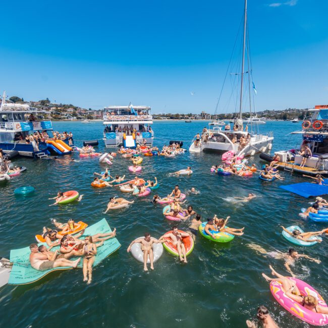 People enjoying a sunny day on the water, floating on colorful inflatables and swimming near several boats. The boats are anchored close together in a picturesque bay with paddleboarders gliding by and houses visible in the background. The water is filled with individuals socializing and relaxing.