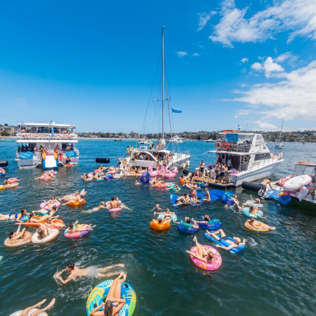 A lively scene of people enjoying a sunny day on the water, with several boats and numerous colorful inflatables. Many are swimming or lounging on the inflatables in a coastal bay, creating a festive atmosphere. A picturesque hilly shoreline and charming houses are visible in the background.