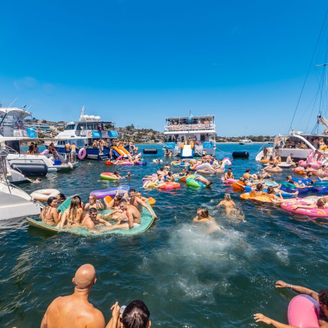 A lively scene of people enjoying a sunny day on boats and in the water, surrounded by inflatables. Several boats, including a large party barge in the background, are anchored closely together. The water is filled with swimmers and colorful kayaks. The sky is clear.