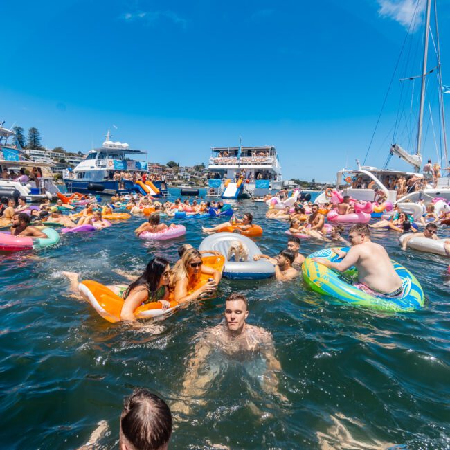 People enjoying a sunny day floating on colorful inflatables and swimming in the water, surrounded by yachts. A festive atmosphere with everyone relaxing and having fun in the bay. A logo reading "Yacht Social Club" is visible in the bottom right corner, highlighting a true marina paradise.