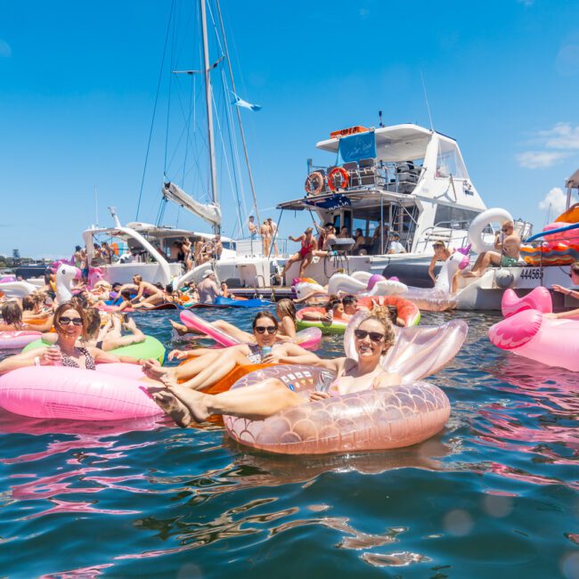 A group of people floating on colorful inflatable loungers and tubes, including a pink flamingo and a unicorn, in a sunny, crowded water area. Several boats are anchored nearby, with people socializing on the decks. The Logo at the bottom right reads "Yacht Social Club," enhancing the festive scene.