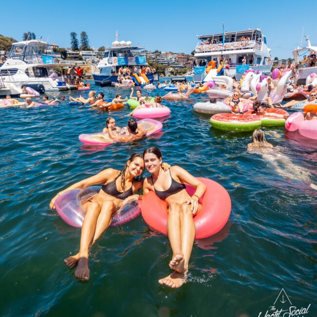 Two women in swimsuits lounge on inflatable rings in a sea crowded with colorful floats and sunbathers. Boats and partygoers visible in the background add to the festive, lively atmosphere, all under "The Yacht Social Club" logo in the bottom right corner.