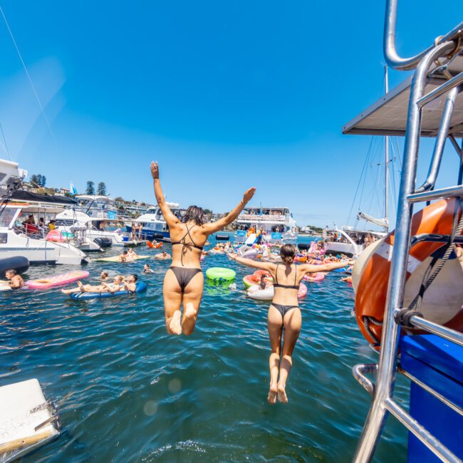 Two women in swimsuits jump into the water from a boat, joining a lively scene of people swimming and floating on inflatables. The sky is clear and blue, with several yachts anchored nearby. A "Yacht Social" logo is visible in the bottom right corner.