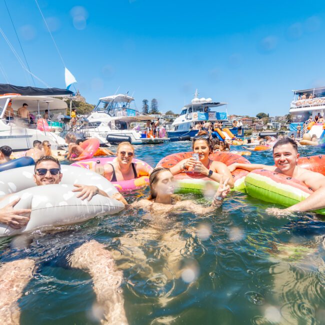 A group of people enjoying a sunny day in the water, floating on inflatable pool toys shaped like donuts and watermelon slices. Behind them, yachts and boats are anchored with others relaxing and socializing. The vibe is lively, festive, and filled with laughter.