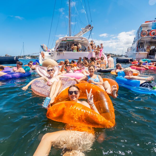 People are enjoying a sunny day on the water, lounging on colorful inflatables near yachts. One person on an orange inflatable kayak makes a peace sign. The scene is cheerful, with clear blue skies, calm water, and various inflatables creating a festive atmosphere.