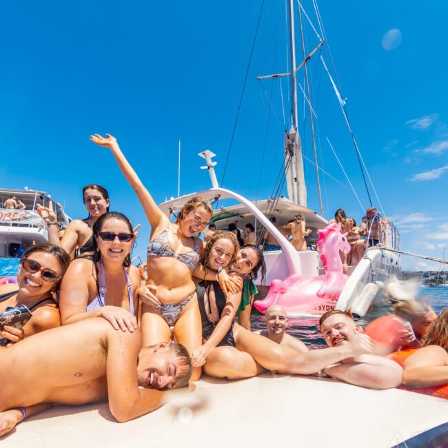 A group of people in swimsuits are enjoying a sunny day on a luxury yacht, smiling and posing for a photo. Some are lying on a floating platform next to the yacht, with inflatable floats and clear blue skies in the background. The atmosphere is cheerful and lively, perfect for relaxation and fun.