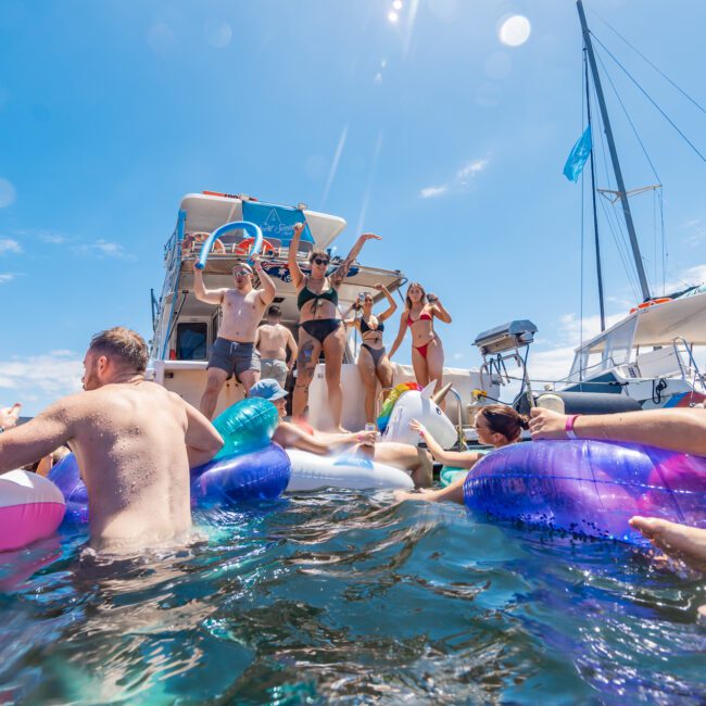 A lively group of people enjoy a sunny day on the water. Some are in colorful inflatable tubes floating near a boat adorned with a flag. More people are on the boat, dancing and having fun while music plays in the background. The sky is clear with a few clouds, enhancing the joyful atmosphere.