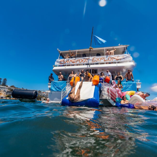 A group of people enjoy a sunny day on and around a large party boat, adorned with banners "Barefoot" and "Explore." Some are lounging on inflatable floats in the water, while others mingle on the boat's deck. A clear blue sky and distant shoreline buildings enhance the cheerful atmosphere.