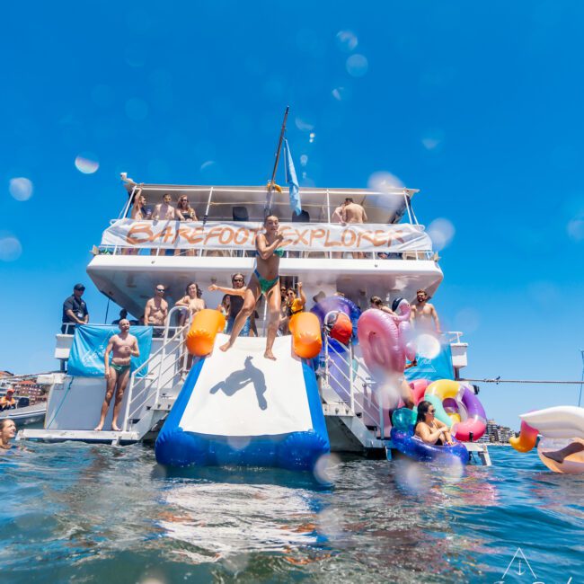 A group of people enjoys a sunny day on a large yacht, some standing on the deck and others in the water. One person slides down an inflatable slide attached to the boat, surrounded by colorful floats. The clear blue sky and vibrant sea create an idyllic backdrop, perfect for this seaside adventure.