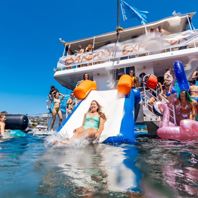 A group of people enjoy a sunny day on a boat party. A woman slides down an inflatable waterslide into the water, surrounded by other people and colorful pool floats. The boat, named "Carefree Express," features a vibrant blue flag fluttering in the breeze.