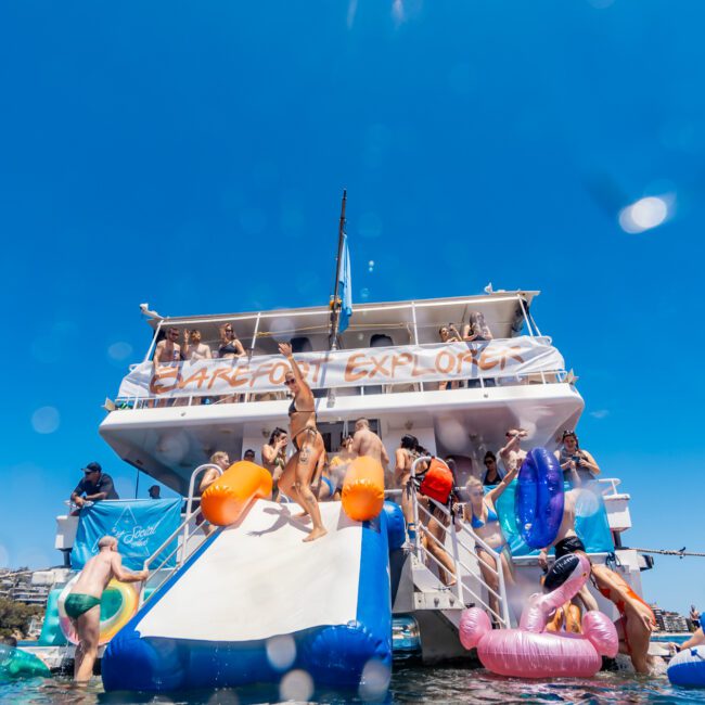 People enjoying a sunny day on a boat named "Barefoot Explorer," some using a blue and white inflatable slide while others float in the water on various inflatables. The boat's deck is filled with more people watching and relaxing under the clear blue sky, making for an unforgettable adventure at sea.