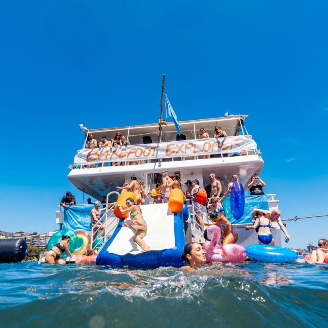 A lively scene on a yacht where several people are enjoying a party. The yacht features a large slide leading to the water. People are swimming and playing with inflatables in the sea. A banner on the yacht reads "Wakepool Explorer" under sunny, clear skies, creating an atmosphere of endless fun and adventure.