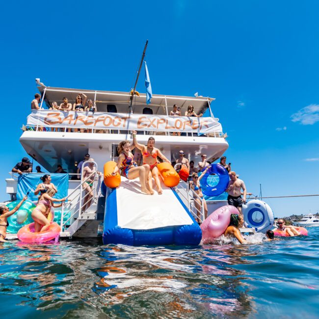 A group of people is enjoying a day out on a large boat, with some on the deck and others swimming. A slide extends from the boat into the water, where several people are using colorful inflatable pool floats. The sky is clear and blue, adding to the perfect atmosphere.