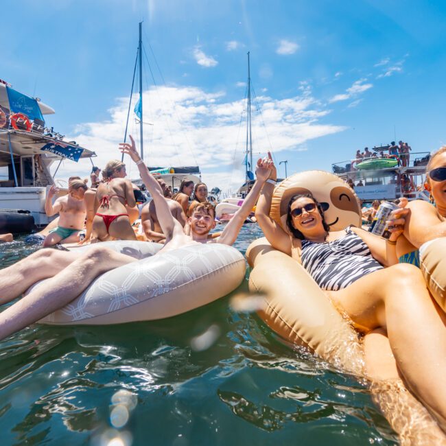 A joyful group of people are relaxing on inflatable floaties in the water next to a boat on a sunny day. Everyone is smiling and holding up their hands joyfully. The clear sky with a few clouds adds to the serene atmosphere, and more people can be seen enjoying themselves on the boat in the background.