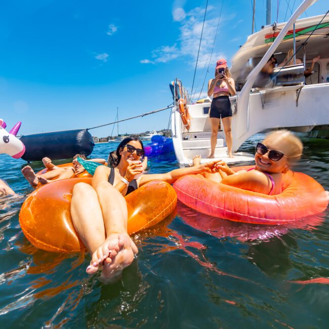 A group of people are relaxing on inflatable floats in the water near a yacht on a sunny day. Two women in sunglasses are smiling and leaning back on their colorful unicorn floats, while others are standing or sitting on the yacht.