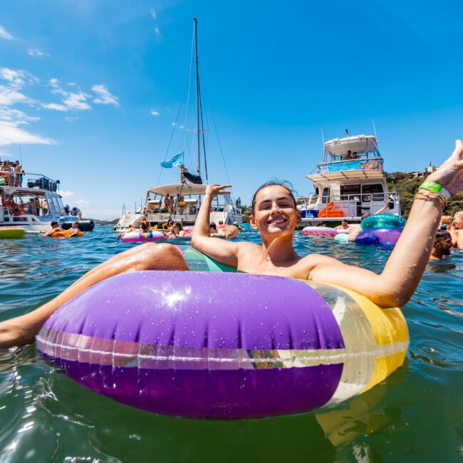 A person is relaxing in a colorful inflatable tube in the water, surrounded by other people on floats. There's a bright, sunny sky and several boats anchored nearby. Hills with greenery create a beautiful backdrop. The atmosphere is lively and joyful, perfect for paddleboarding enthusiasts.