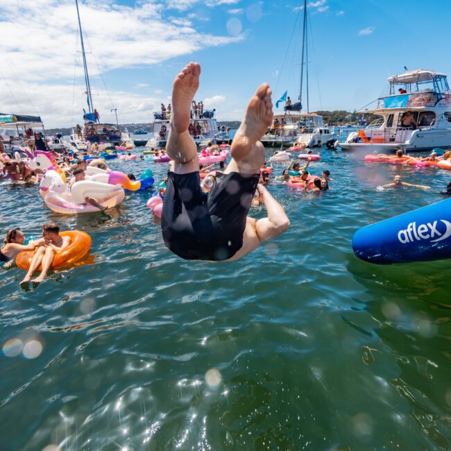 A person mid-air diving into the water surrounded by people relaxing on inflatable floats during a sunny day at a lively boat party. Various boats and yachts are anchored in the background, with blue skies and scattered clouds overhead, creating an idyllic summer setting.