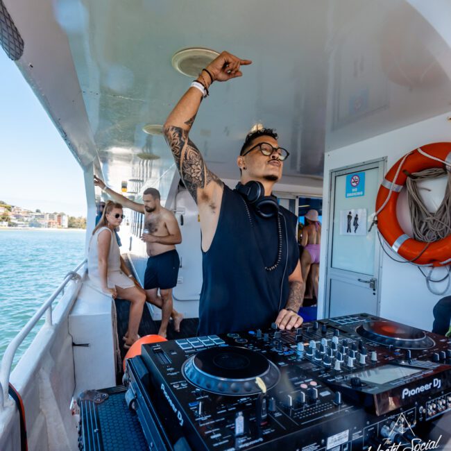 A DJ wearing a sleeveless black shirt and headphones is performing on a yacht with a mixer and turntables. Behind him, people are enjoying the sunny day on the deck, with two individuals in swimsuits standing nearby. Palm trees sway gently in the background.