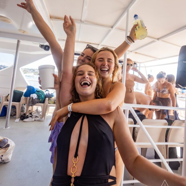 Three women, smiling and posing energetically on a yacht. The woman in the foreground wears a black swimsuit and is being hugged from behind by another woman. People, various beach items, and vibrant summer accessories are visible on the boat's deck. The sun is shining brightly.