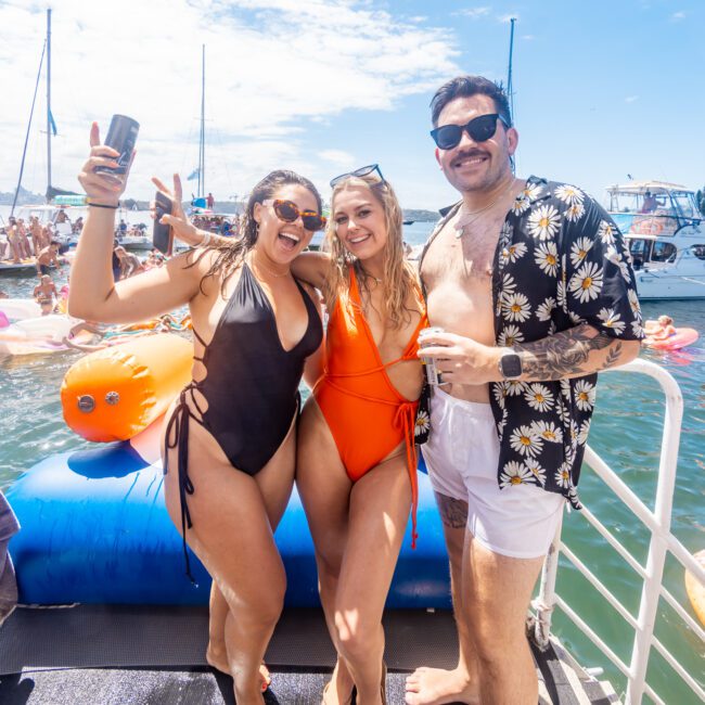 Three people are standing on a dock or boat in swimwear. Two women in black and orange swimsuits hold drinks and smile at the camera, while a man in a floral shirt, white shorts, and sunglasses stands next to them. Boats and floaties are visible in the background, capturing the essence of The Yacht Social Club Sydney Boat Hire.