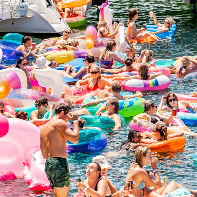 A lively scene of numerous people enjoying a sunny day on the water, floating on colorful inflatable rafts, including unicorns and donuts. Some are swimming, others are sunbathing, and everyone seems to be having a great time. Luxury Yacht Rentals Sydney offers an ideal backdrop with boats docked in the background.