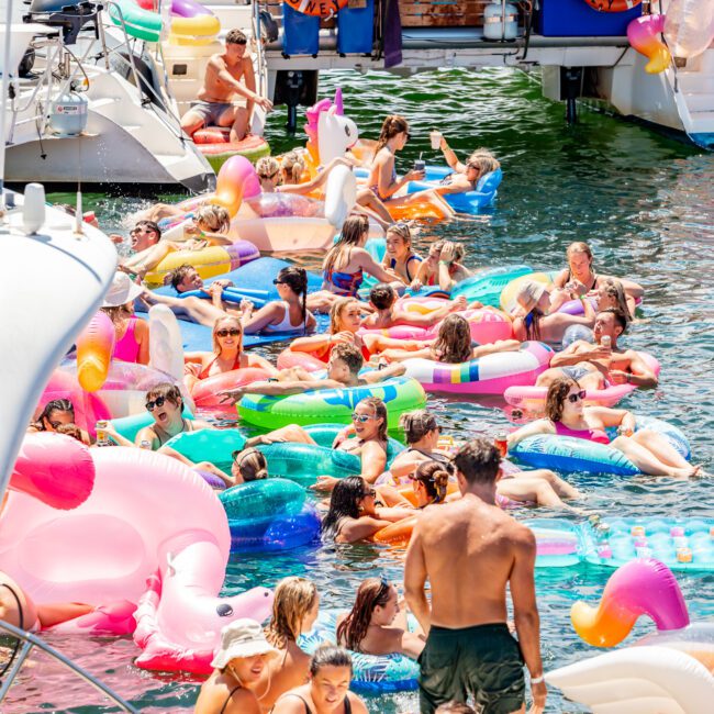 A large group of people enjoying a sunny day on the water, floating on colorful inflatables like unicorns and pink flamingos. Luxury yachts from The Yacht Social Club are docked nearby, and people are socializing, swimming, and lounging in swimsuits. The atmosphere is lively and festive.