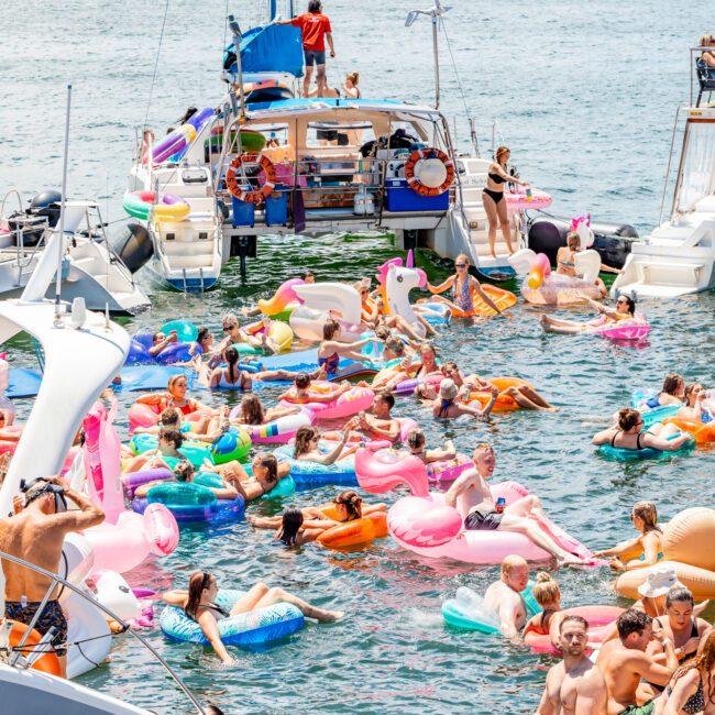 A lively scene of people enjoying a sunny day on a lake, surrounded by various boats. The water is filled with colorful inflatable pool floats, including flamingos, swans, and donuts. Dozens of people are relaxing and socializing, creating a vibrant atmosphere reminiscent of The Yacht Social Club Event Boat Charters.