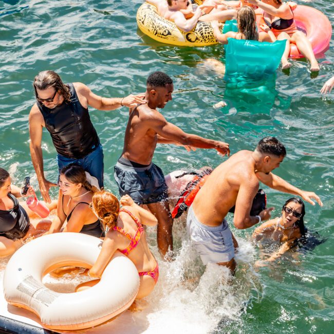 A lively scene with many people enjoying a sunny day on the water using various inflatable pool floats. Some are standing or sitting on the floats, while others swim and dance. The water is greenish, and the mood is festive. A watermark reads "Sydney Harbour Boat Hire The Yacht Social Club.