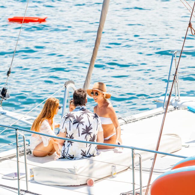 Three people are relaxing and conversing on the deck of a sailboat. The man is wearing a palm tree-patterned shirt, one woman is in a white bikini and sunhat, and the other woman is also in a white bikini. The Yacht Social Club Sydney Boat Hire ensures the boat is docked on calm, blue water under sunny skies.