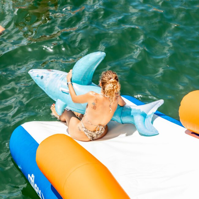 A person with wet hair and a tattoo on their back sits on an inflatable raft, holding a large inflatable shark. The raft is floating in green water near another inflatable toy with a unicorn head. Experience more water fun with The Yacht Social Club Sydney Boat Hire for unforgettable adventures!