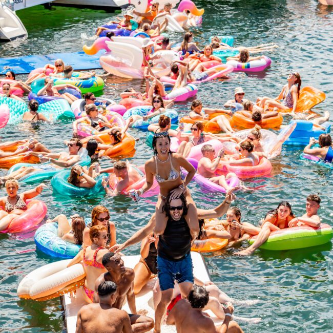 A lively scene on the water with many people enjoying a summer day. Guests are floating on colorful inflatables, swimming, and socializing. Two boats in the background are filled with more people, while some are dancing on a floating platform in the center. It's a perfect day for boat parties with The Yacht Social Club.