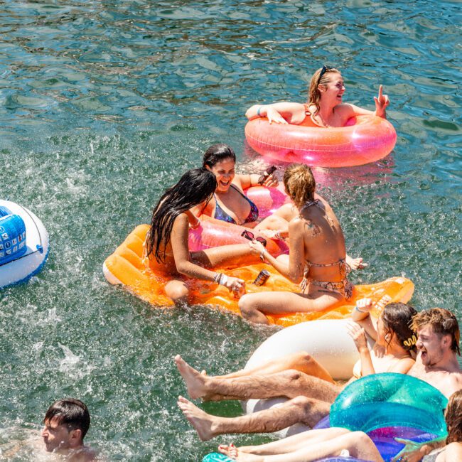 A group of people enjoying a sunny day in the water with various inflatable floats. They appear to be laughing and interacting, creating a lively atmosphere. Some of the floats include a pink ring, an orange mattress, and a colorful lounge at The Yacht Social Club Event Boat Charters.