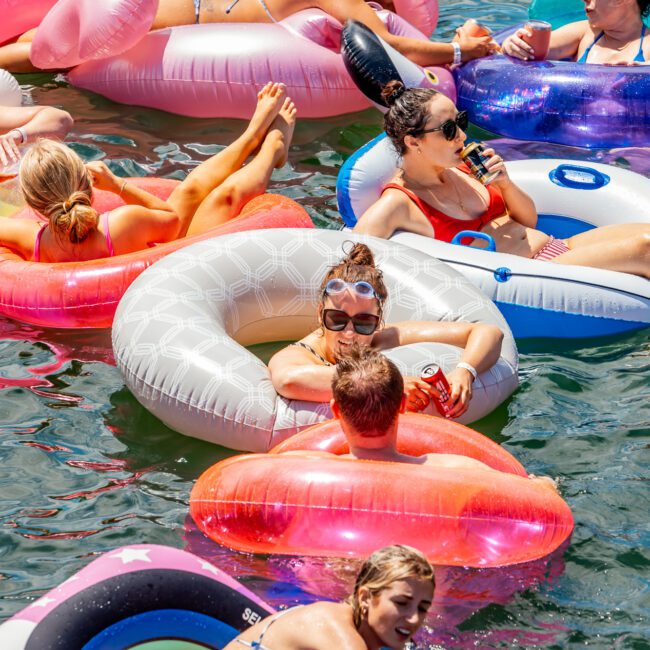 A group of people relaxing on colorful inflatable pool floats in a body of water under bright sunlight. Most are wearing swimsuits and sunglasses, chatting and enjoying beverages. The overall mood is vibrant and lively, much like the atmosphere at Boat Parties Sydney The Yacht Social Club.