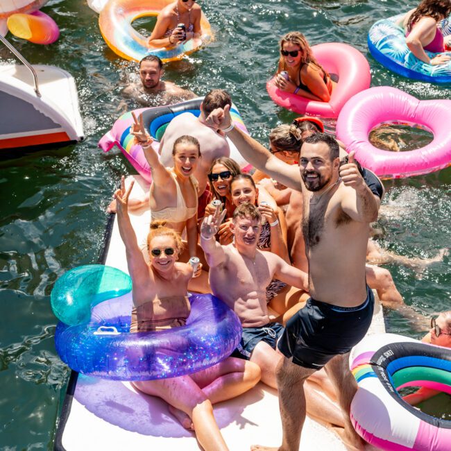 A group of people enjoying a sunny day on the water, sitting on an inflatable platform surrounded by colorful floaties. They are smiling, posing for the camera, and making peace signs. Nearby, others swim and float in the clear, inviting water—courtesy of Sydney Harbour Boat Hire The Yacht Social Club.