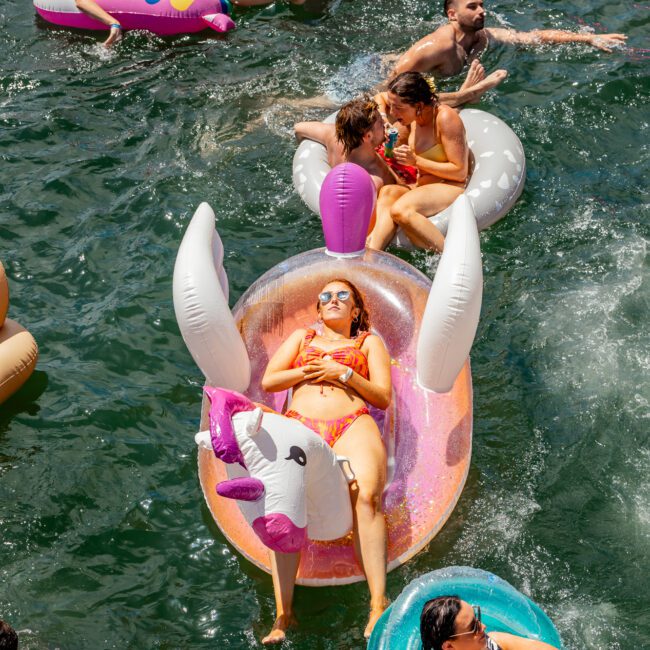 A group of people floats on various inflatable pool toys in a body of water during The Yacht Social Club Event. A woman wearing sunglasses and a yellow bikini relaxes on a unicorn float in the center, surrounded by others on flamingo, pineapple, and doughnut floats. It appears to be a sunny day.