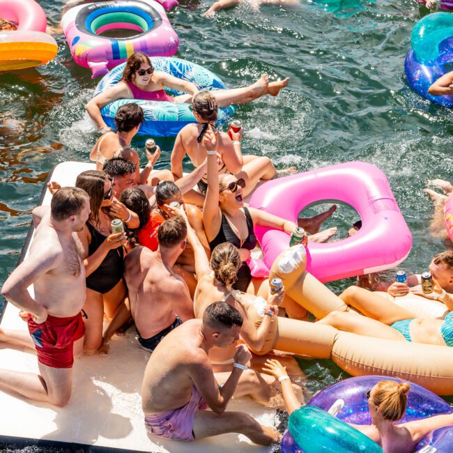 A group of people in swimwear enjoys a sunny day on a lake, floating on colorful inflatable rafts and rings. They are laughing, relaxing, and holding drinks while socializing. The water is calm, and everyone appears to be having a great time—just like at The Yacht Social Club Sydney Boat Hire event.