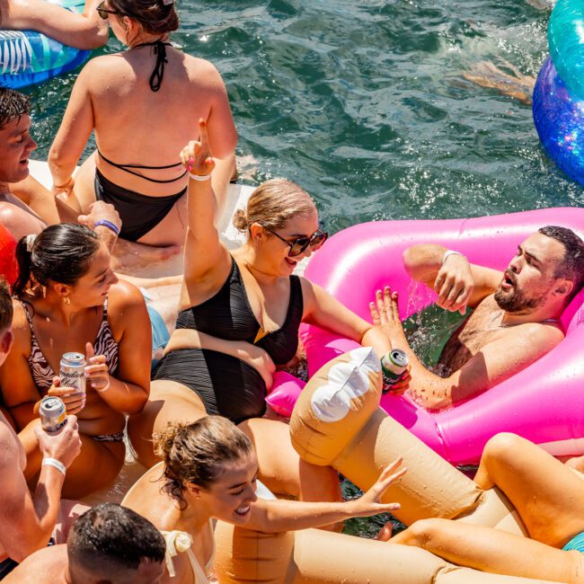 A group of people are having fun on inflatable floats in the water near a boat rental from The Yacht Social Club event. They are swimming, drinking from cans, and socializing. One person is making a peace sign while others smile and talk. The scene is lively and energetic, with bright sunlight illuminating the gathering.