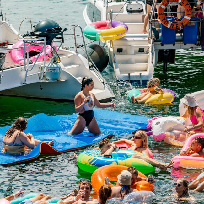 People float on colorful inflatables in the water near two docked boats. Some are on large blue mats while others are on various floaties, enjoying a sunny day. The boats, part of The Yacht Social Club Sydney Boat Hire, are adorned with more inflatables and a life ring. It's a lively, festive scene.