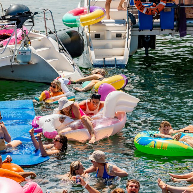 A lively scene at a waterfront, with numerous people enjoying the water on colorful inflatables. The crowd features adults and children on unicorn, flamingo, and various other floaties near docked boats, soaking up the sun and having fun at The Yacht Social Club Event Boat Charters.