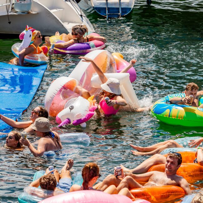 A lively scene with people enjoying a day on the water, floating on various inflatable pool toys and lounging near boats. The water is filled with colorful inflatables, including a unicorn and flamingo, as everyone relaxes and mingles under the sun. It's reminiscent of a Boat Parties Sydney The Yacht Social Club event.