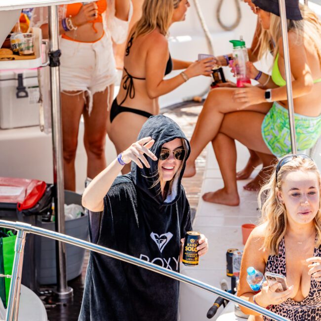 A group of people on a boat enjoying a sunny day. One person in the foreground is wearing a black hooded towel with sunglasses, smiling, and holding a drink. Others in the background are sitting and chatting, dressed in swimwear. Experience this with The Yacht Social Club Sydney Boat Hire for unforgettable moments.