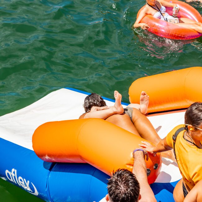 A group of people enjoying a sunny day on the water with The Yacht Social Club. One person slides down an inflatable water slide into the lake, while others float nearby on orange and pink inner tubes. The atmosphere is lively and fun, capturing the essence of Boat Parties Sydney The Yacht Social Club.