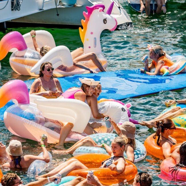 A group of people relaxing on colorful inflatable floats, including a unicorn, swan, and donut, in the water next to a boat from The Yacht Social Club Sydney Boat Hire. They are enjoying sunny weather in swimsuits and hats, with some holding drinks. The atmosphere appears lively and festive.