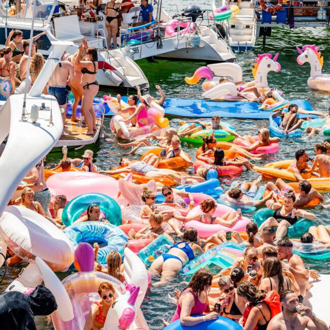 A lively scene at a lake party with numerous people on inflatable pool floats, including unicorns and donuts, gathered around several boats. Attendees are socializing, sunbathing, and enjoying the water in colorful attire under a bright, sunny sky—a perfect setting for The Yacht Social Club Event Boat Charters.