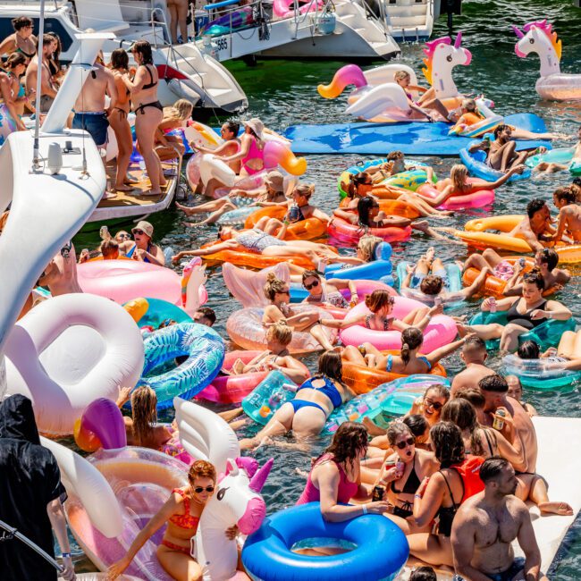 A large group of people enjoying a sunny day on the water with many colorful inflatable floats, including unicorns and flamingos. Boats are docked nearby, and people are swimming, lounging, and socializing on the inflatables at The Yacht Social Club Sydney Boat Hire event.