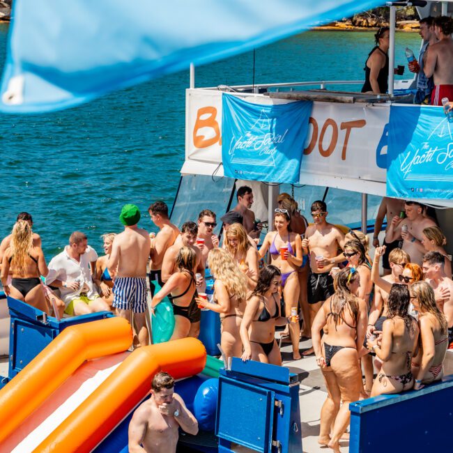 A lively scene on a party boat from The Yacht Social Club Sydney Boat Hire shows numerous people in swimsuits enjoying the sun and water activities. An inflatable slide leads into the water, where a person is climbing out. The boat is decorated with banners, and the water surrounding is clear and blue.