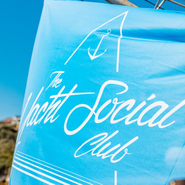 A bright blue flag with white text that reads "The Yacht Social Club" and features an illustration of a yacht. The flag is attached to a pole with a clear blue sky in the background, highlighting the luxury yacht rentals Sydney offers. The Instagram handle "@theyachtsocialclub" is also visible at the bottom-left corner.