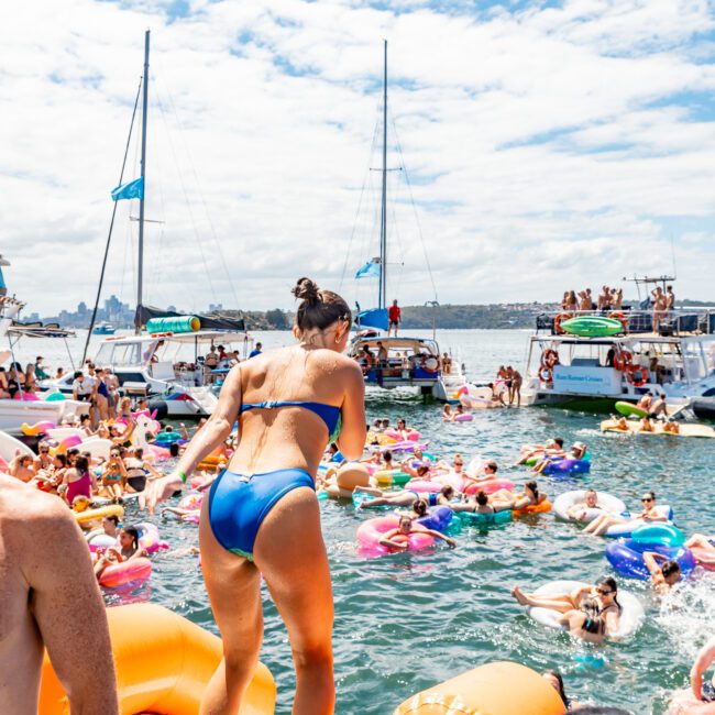 A woman in a blue bikini stands on the edge of an inflatable structure, about to jump into a crowded, lively pool. The pool is filled with people enjoying themselves and floaties under a clear sky; yachts, including those from Luxury Yacht Rentals Sydney, are visible in the background.